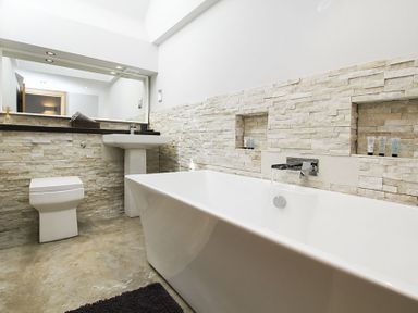 A bathroom with a bathtub, pedestal sink, toilet, stone wall tiles, and a large mirror at Rhyd Angharad Barns in Rhydwyn
