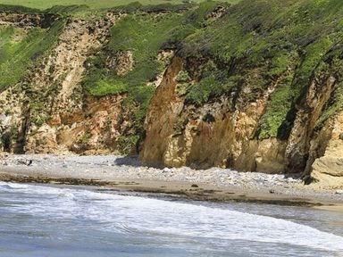 A rocky cliff with green vegetation above a pebble and sandy beach with waves at Rhyd Angharad Barns in Rhydwyn