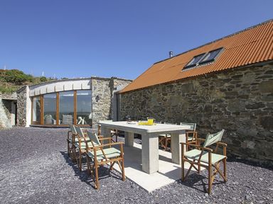 An outdoor seating area with a stone table and eight chairs next to a stone building with a corrugated orange roof at Rhyd Angharad Barns in Rhydwyn