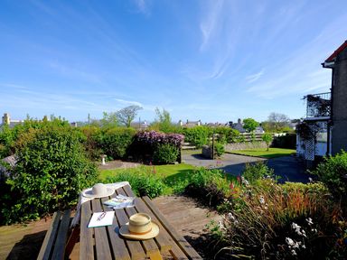 An outdoor wooden picnic table with two hats magazines and a notebook in a garden with shrubs flowers and houses in the background at Seiriol View in Moelfre