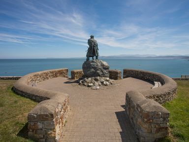 A statue of a person standing on a rock facing the sea with stone walls and paved path around it at Seiriol View in Moelfre