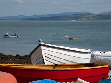 A beach scene with a red wooden boat and a white rowboat with other boats in the water and hills in the background at Seiriol View in Moelfre
