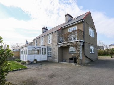 A two-story stone house with a white conservatory and a balcony with climbing plants and outdoor seating at Seiriol View in Moelfre