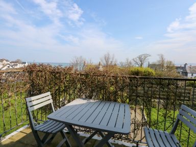 A balcony with two chairs and a table overlooking a garden and houses with the sea in the background at Seiriol View in Moelfre