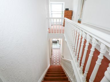 A staircase with red carpet and white banisters leading to a hallway with patterned floor tiles and a wooden dresser at Seiriol View in Moelfre