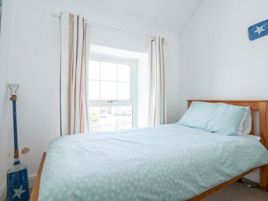 A bedroom with a bed with light blue bedding near a window with striped curtains and a decorative paddle beside the bed at Seiriol View in Moelfre