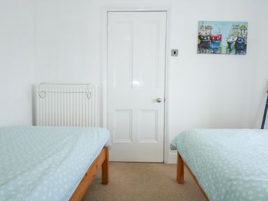 A bedroom with two beds covered in light blue bedspreads a white door a radiator and a painting of boats at Seiriol View in Moelfre