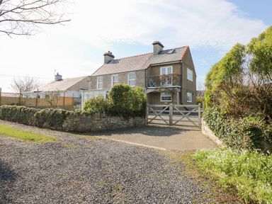 A stone and brick house with a tiled roof behind a wooden gate with gravel and greenery around at Seiriol View in Moelfre