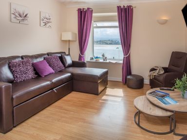 A living room with a brown leather sectional sofa with purple cushions a round wooden coffee table with magazines a brown armchair a window with purple curtains and a view of water at The Mermaid in Brynsiencyn
