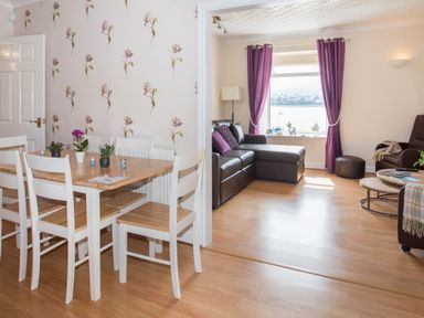 A dining area with a wooden table and white chairs next to a living room with a brown leather sectional sofa and purple curtains at The Mermaid in Brynsiencyn