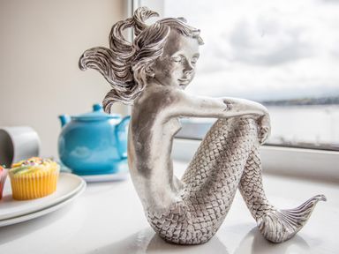 A silver mermaid figurine sitting on a windowsill with a blue teapot and cupcakes on a plate nearby at The Mermaid in Brynsiencyn