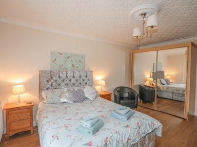 A bedroom with a bed with floral bedding two wooden bedside tables lamps a black armchair and a mirrored wardrobe at The Mermaid in Brynsiencyn