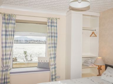 A bedroom with a window seat with cushions overlooking water and an open white wardrobe with towels and a lamp at The Mermaid in Brynsiencyn