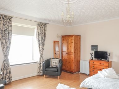 A bedroom with wooden wardrobe and chest of drawers a black armchair and window with floral curtains at The Mermaid in Brynsiencyn