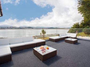 An outdoor patio with rattan furniture and white cushions overlooking a body of water at The Mermaid in Brynsiencyn