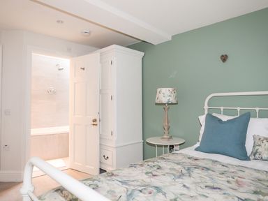 A bedroom with a bed with floral bedding a blue pillow a side table with a lamp a white wardrobe and an open door showing a bathroom at The Honey Pot in Garndolbenmaen