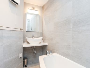 A bathroom with a wall-mounted sink basin a mirror a bathtub and gray tiled walls at The Honey Pot in Garndolbenmaen