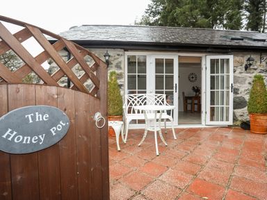 An outdoor patio area with a wooden gate labeled The Honey Pot white metal table and chairs and open double doors leading inside at The Honey Pot in Garndolbenmaen