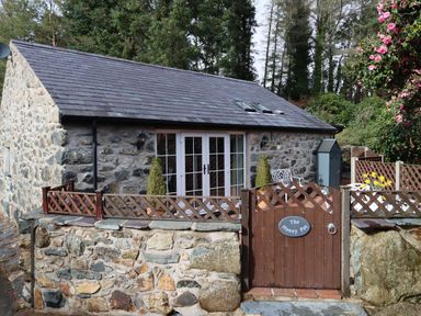 A stone cottage with a wooden gate and fence surrounded by trees and flowers at The Honey Pot in Garndolbenmaen