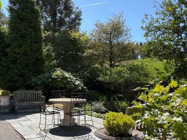 An outdoor patio with a round stone table and metal chairs surrounded by trees and bushes at The Honey Pot in Garndolbenmaen