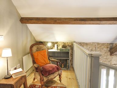 A chair with cushions next to a small table with a lamp and an open book on a wooden floor near a stone wall at Three Gates in Llangoed