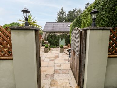 An open wooden gate leading to a stone patio with potted plants a small building with a green door and outdoor seating surrounded by trees at The Little Coach House in Garndolbenmaen