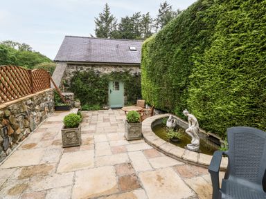 An outdoor patio area with stone flooring a stone building with a green door a hedge two planters a stone fountain with statues a round wooden table with chairs and a plastic chair at The Little Coach House in Garndolbenmaen