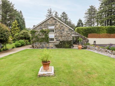 A stone house with two windows surrounded by trees and grass with potted plants in the yard at The Little Coach House in Garndolbenmaen