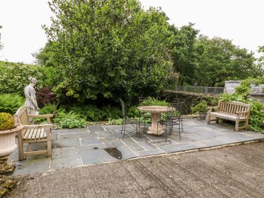 A garden patio with two wooden benches metal chairs a round stone table plants and a statue at The Little Coach House in Garndolbenmaen