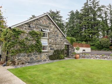 A stone house with climbing plants and a lawn in front surrounded by trees and a paved pathway at The Little Coach House in Garndolbenmaen