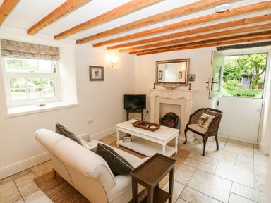 A living room with a beige sofa a white coffee table a wooden chair a small TV a fireplace and an open door showing a garden at The Little Coach House in Garndolbenmaen