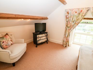 A room with a beige armchair a small black dresser with a television and floral curtains at The Little Coach House in Garndolbenmaen
