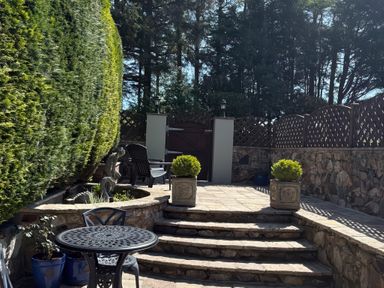 An outdoor stone patio with steps leading to a gate metal table and chairs stone planter boxes and a wooden fence at The Little Coach House in Garndolbenmaen