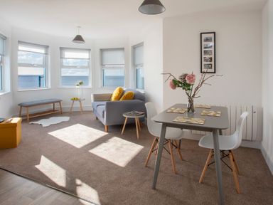 A living room with a gray sofa and yellow cushions a small dining table with two white chairs flowers in a vase a bench and windows showing the sea at The Towers - Ardudwy in Criccieth