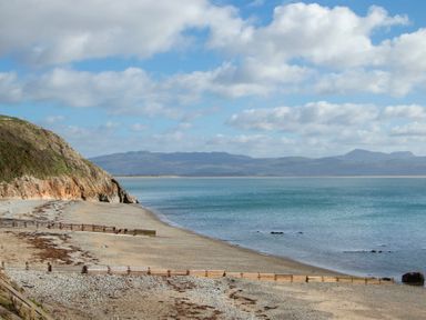 A beach with a rocky cliff on the left and wooden groynes along the shore with mountains in the background at The Towers - Ardudwy in Criccieth
