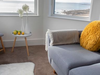 A corner of a room with a gray sofa with a yellow pillow and a blanket next to a small table with vases and fruit by two windows overlooking a rocky beach at The Towers - Ardudwy in Criccieth
