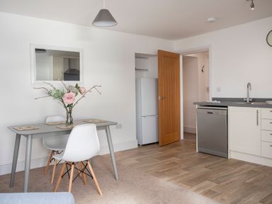 A dining area with a grey table and white chairs next to a kitchen with white cabinets and a dishwasher at The Towers - Ardudwy in Criccieth