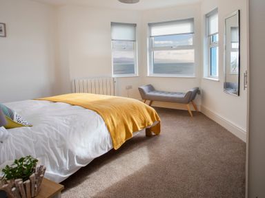 A bedroom with a bed and yellow blanket and a grey bench by the windows overlooking the sea at The Towers - Ardudwy in Criccieth