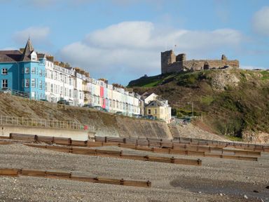 A coastal scene with colorful row houses along a cliff and a castle ruin on a hill at The Towers - Ardudwy in Criccieth