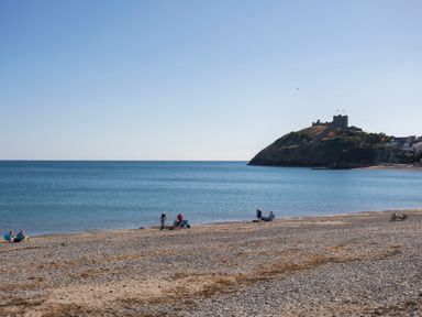 A pebble beach with people sitting by the water and a hill with a castle in the background at The Towers - Ardudwy in Criccieth