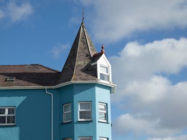 A blue building with a pointed turret and white windows against a cloudy sky at The Towers - Castell in Criccieth