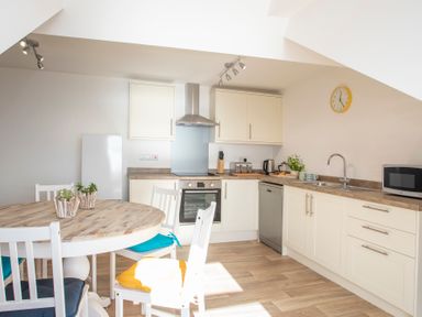 A kitchen with white cabinets, a round wooden dining table with chairs, a stove, dishwasher, microwave, and a wall clock at The Towers - Castell in Criccieth