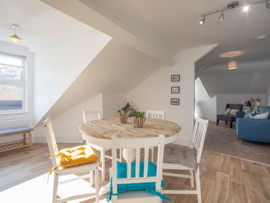 A dining area with a round wooden table and white chairs with cushions next to a window overlooking the sea at The Towers - Castell in Criccieth