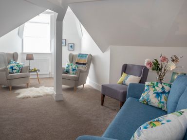 A living room with blue and beige chairs a blue sofa with floral cushions and a small table with a lamp and fruit at The Towers - Castell in Criccieth