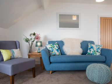 A living room with a blue sofa and a grey armchair with cushions a wooden side table with a lamp and flowers a knitted pouf and a wall mirror at The Towers - Castell in Criccieth