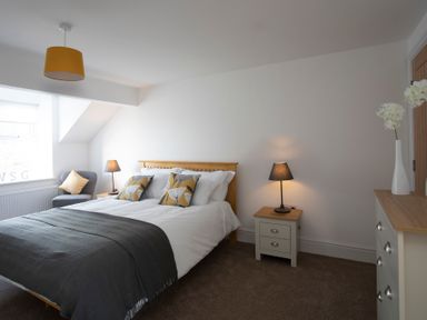 A bedroom with a bed with pillows and throws two bedside tables with lamps a chair with a cushion a chest of drawers with a vase of flowers and a ceiling light at The Towers - Castell in Criccieth