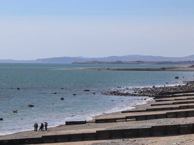 A rocky beach with wooden groynes and people standing near the water with hills in the distance at The Towers - Castell in Criccieth