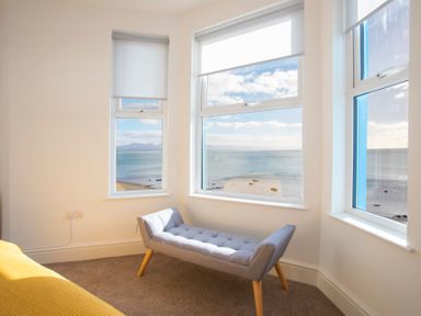 A corner room with three windows overlooking the sea and a gray bench with wooden legs at The Towers - Llyn in Criccieth
