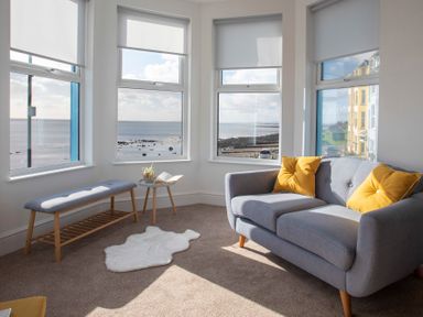 A living room with a gray sofa and yellow cushions near windows overlooking the sea at The Towers - Llyn in Criccieth