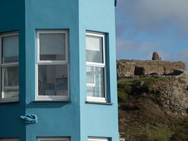 A corner of a blue building with white framed windows and a stone ruin on a hill in the background at The Towers - Llyn in Criccieth